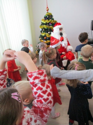Mikołaj siedzi na krześle i mówi do mikrofonu. Dzieci tańczą.
